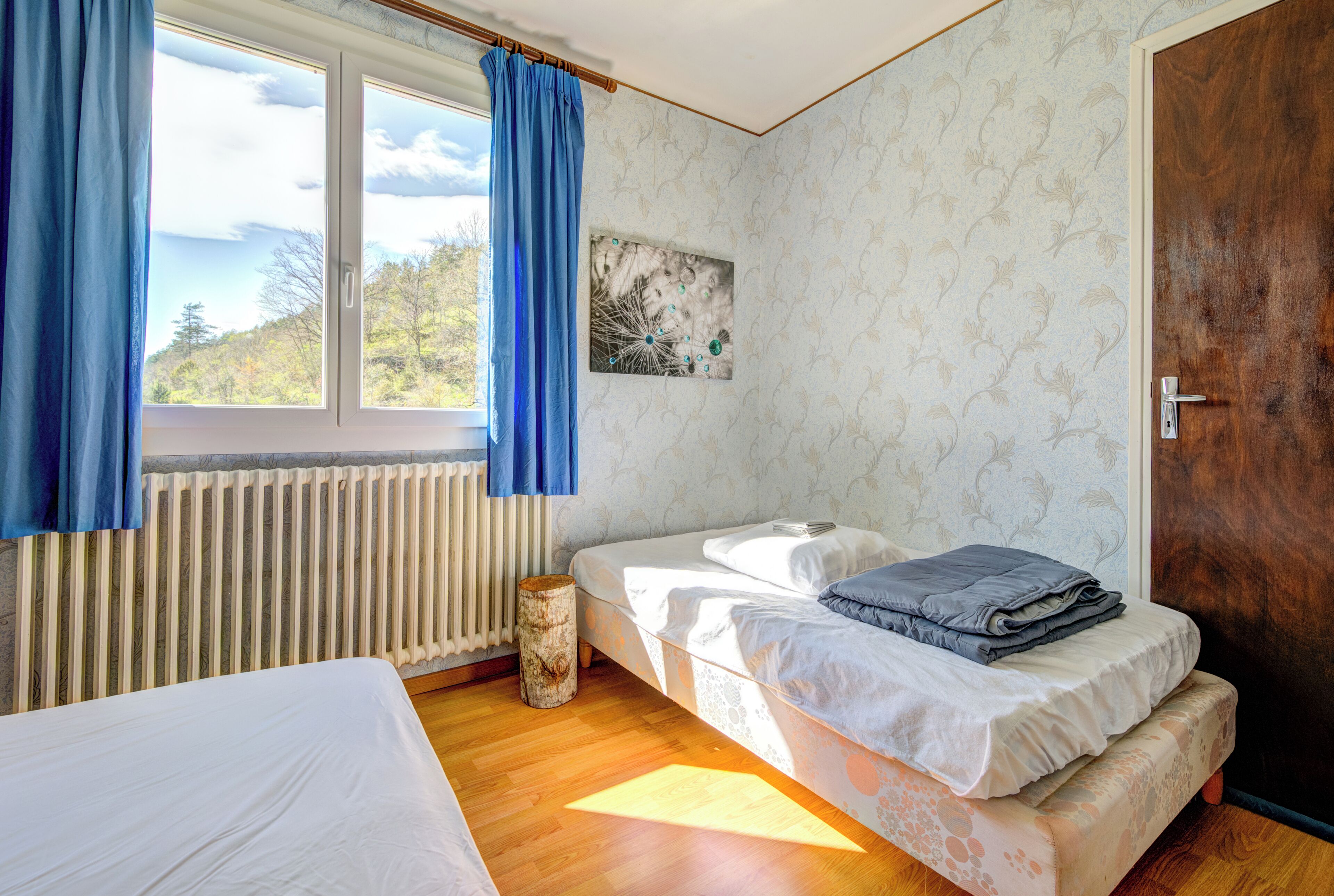 Ein Schlafzimmer mit zwei Betten, einem Fenster mit blauen Vorhängen, einem Heizkörper und einem Holzboden.