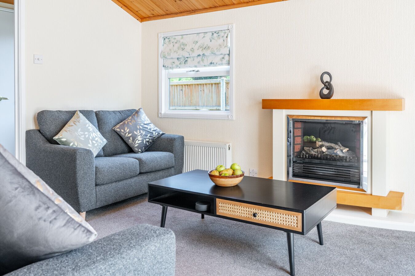A living room with a grey sofa, a black coffee table with a woven basket, and a fireplace with logs.