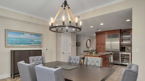 A dining area with a large table, six grey upholstered chairs, a chandelier, a painting of a beach, and a kitchen with modern appliances.