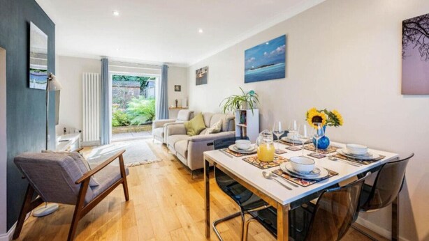 Un comedor moderno con una mesa de madera dispuesta para una comida, un sofá y vistas a un jardín a través de un ventanal.