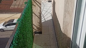 A narrow balcony with a tiled floor, a green hedge, and a view of neighboring buildings and a clear sky.
