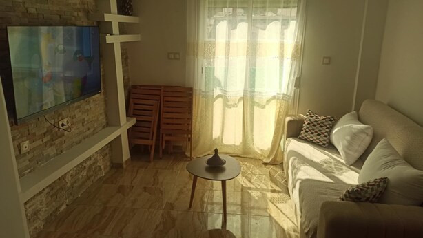 A living room with a TV mounted on a brick wall, a sofa with patterned cushions, a small table, and a window with sheer curtains.
