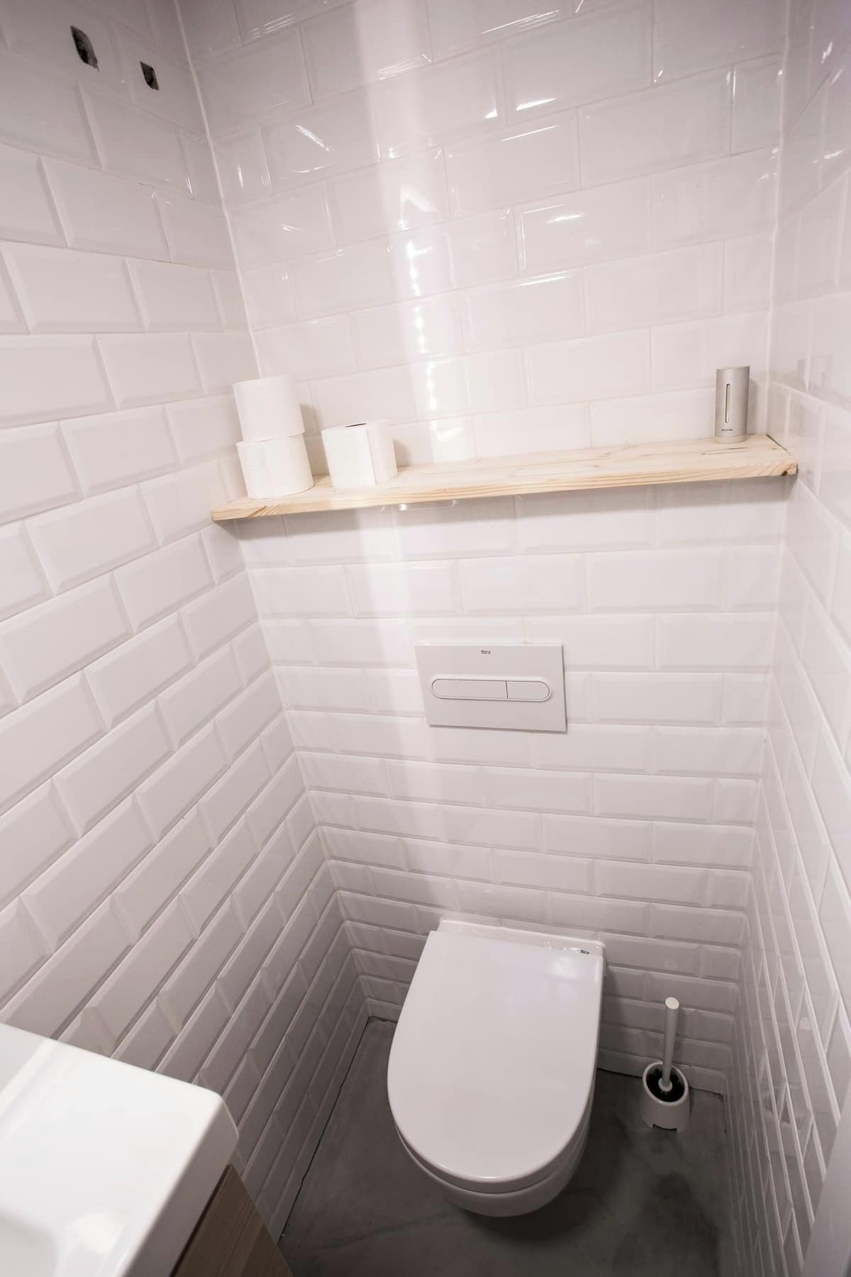 Un baño pequeño con azulejos blancos, un inodoro, un estante con papel higiénico y un secamanos, y un tirador de la cisterna en la pared.