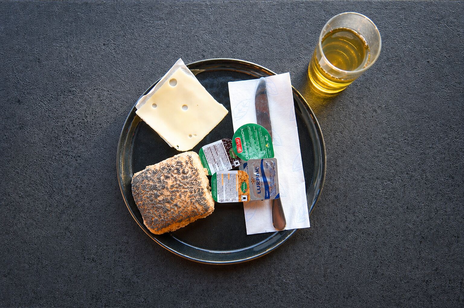 A plate with a slice of cheese, a piece of bread, a glass of beer, and two packaged snacks.