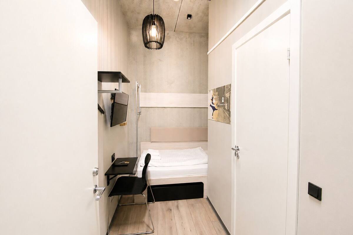 A compact hotel room with a bed, a desk, a chair, a TV, and a pendant light.