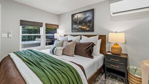 Une chambre moderne avec un grand lit, une table de chevet avec une lampe, un tableau au mur et une vue sur un jacuzzi situé à l’extérieur.