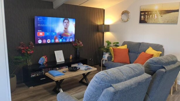 Una sala de estar moderna con un televisor de pantalla plana, un sofá gris con cojines naranja y amarillos, una mesa de centro de madera y un panel decorativo en la pared.