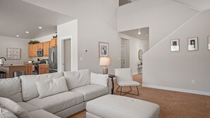 A modern living room with a sectional sofa, a small table, and a chair. There is a kitchen in the background with wooden cabinets and a refrigerator. - Wasatch Peak Chateau R3 | Ski Snowbasin | Hot Tub (Mountain Green)