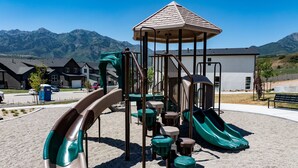 A playground with slides and climbing structures, set against a backdrop of mountains and residential buildings.