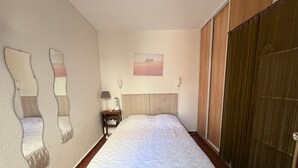 A hotel room with a large bed, a bedside table with a lamp, a wall-mounted mirror, and a painting above the bed. - Renovated 2BR Ground Floor, 4P, Pets OK, Parking, 400m Beach (Le Barcarès)