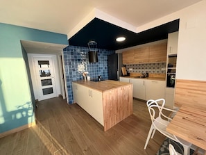 Cozinha moderna com ilha de madeira, painel de parede azul e área de refeições com uma cadeira branca.