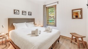 En pænt redt seng med hvide sengetøj og håndklæder, et sengebord med en lampe, en træskammel, et stort vindue med udsigt til grøn vegetation, og tre billeder hængende på væggen.