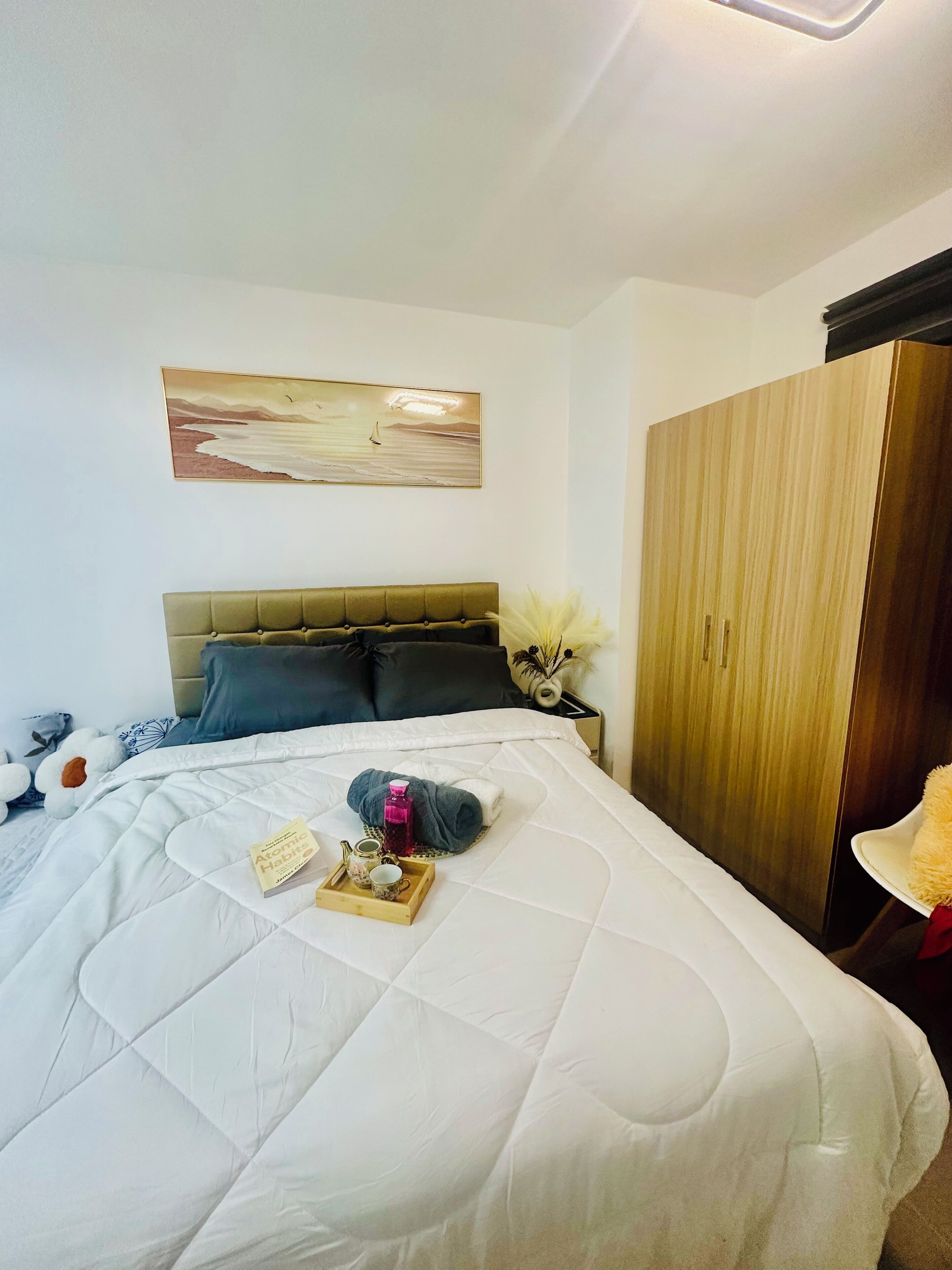 A neatly made bed with a white duvet, a bedside table with a vase and a bottle, and a painting above the bed.