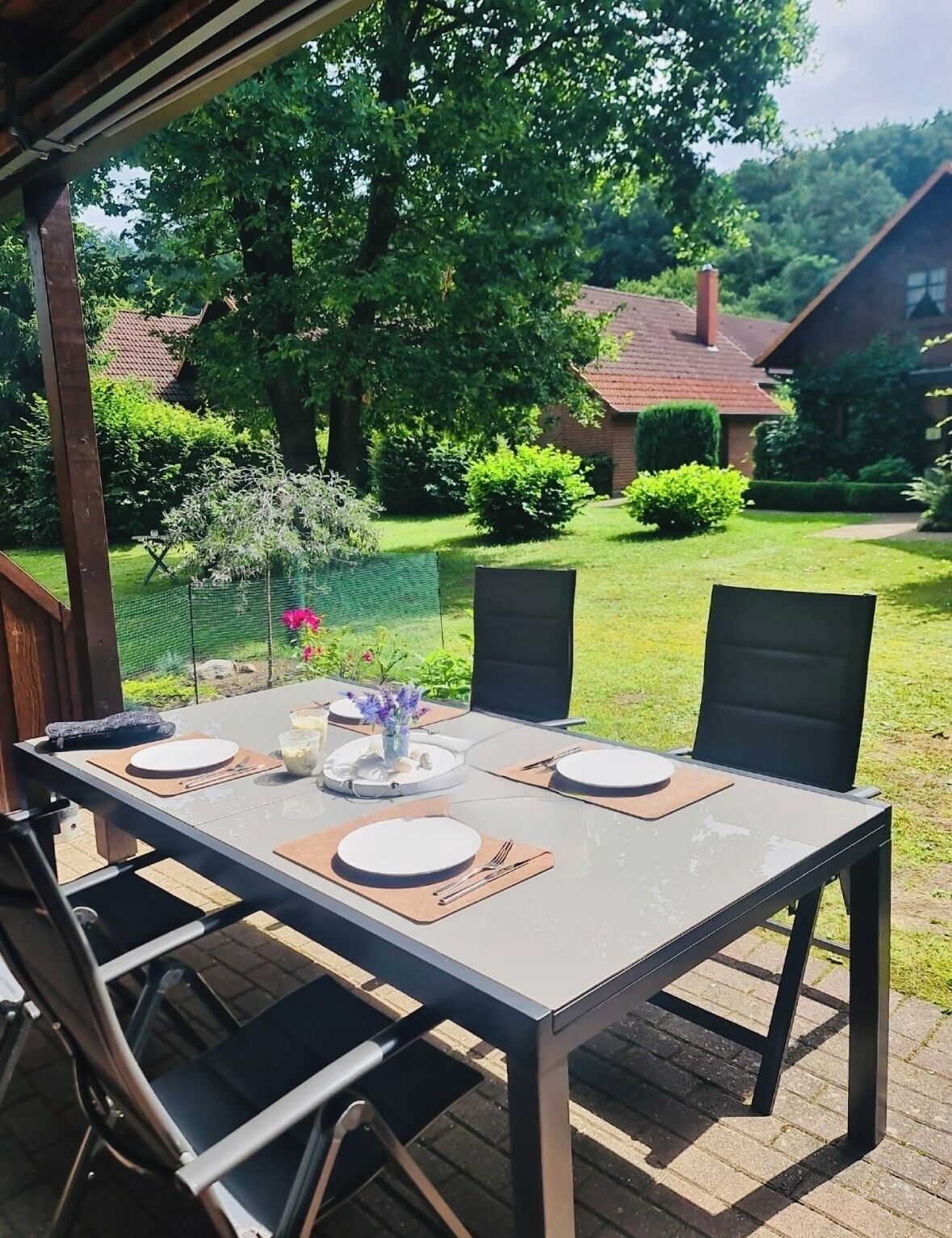 Ein Essbereich im Freien mit einem Tisch für vier Personen, umgeben von Stühlen, in einem Garten mit einem Haus und Bäumen im Hintergrund.