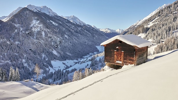 A wooden cabin with a snow-covered roof, surrounded by snow-covered mountains and trees.