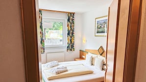 A bedroom with a wooden bed, white linens, a view of greenery outside, a radiator, and a framed picture on the wall.