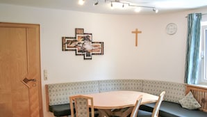 A dining area with a round table, four chairs, a bench, a wooden door, a wall-mounted cross, and a clock.