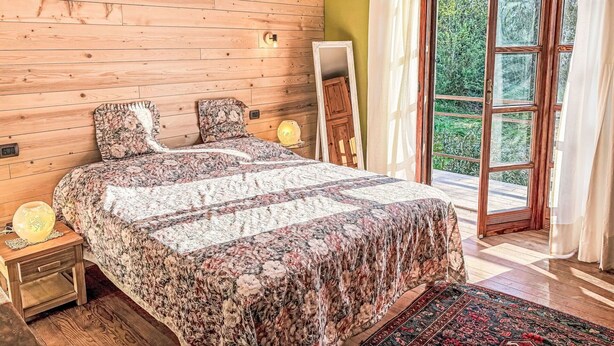 A bedroom with a wooden slat wall, a bed with floral bedding, a bedside lamp, and a view through a glass door to an outdoor area. - Wonderful private villa for 6 guests with WIFI, A/C, TV and terrace (Asti)