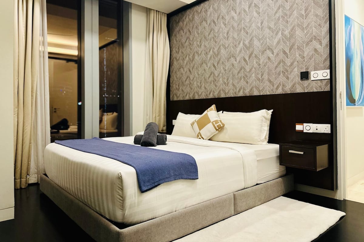 Ein modernes Hotelzimmer mit einem großen Bett, einem geometrisch gemusterten Kopfteil, einem Nachttisch und Blick auf einen Schrank mit Kleidung.