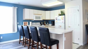A modern kitchen with white cabinets, a central island with bar stools, a microwave, a coffee maker, and a refrigerator. - Beautiful 3 Bd condo on 23rd Street with outdoor pool. (Ocean City)