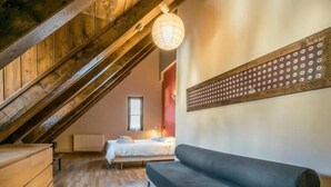 Ein gemütliches Schlafzimmer im Loft-Stil mit schrägen Wänden, Holzbalken, einem großen Fenster, einem Bett, einer Couch und einer Kommode.