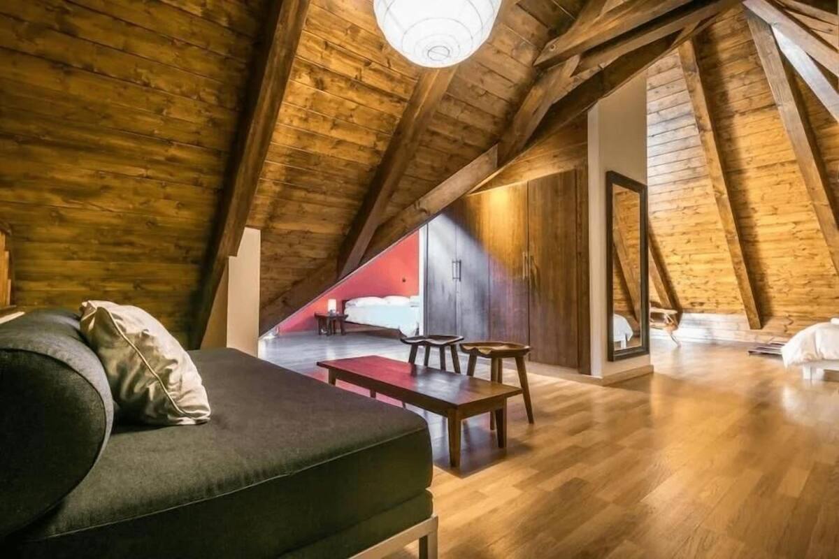 Une chambre mansardée chaleureuse avec un plafond en pente, des poutres en bois, un canapé vert, une petite table à manger et une vue sur une pièce rouge, visible à travers une grande fenêtre.
