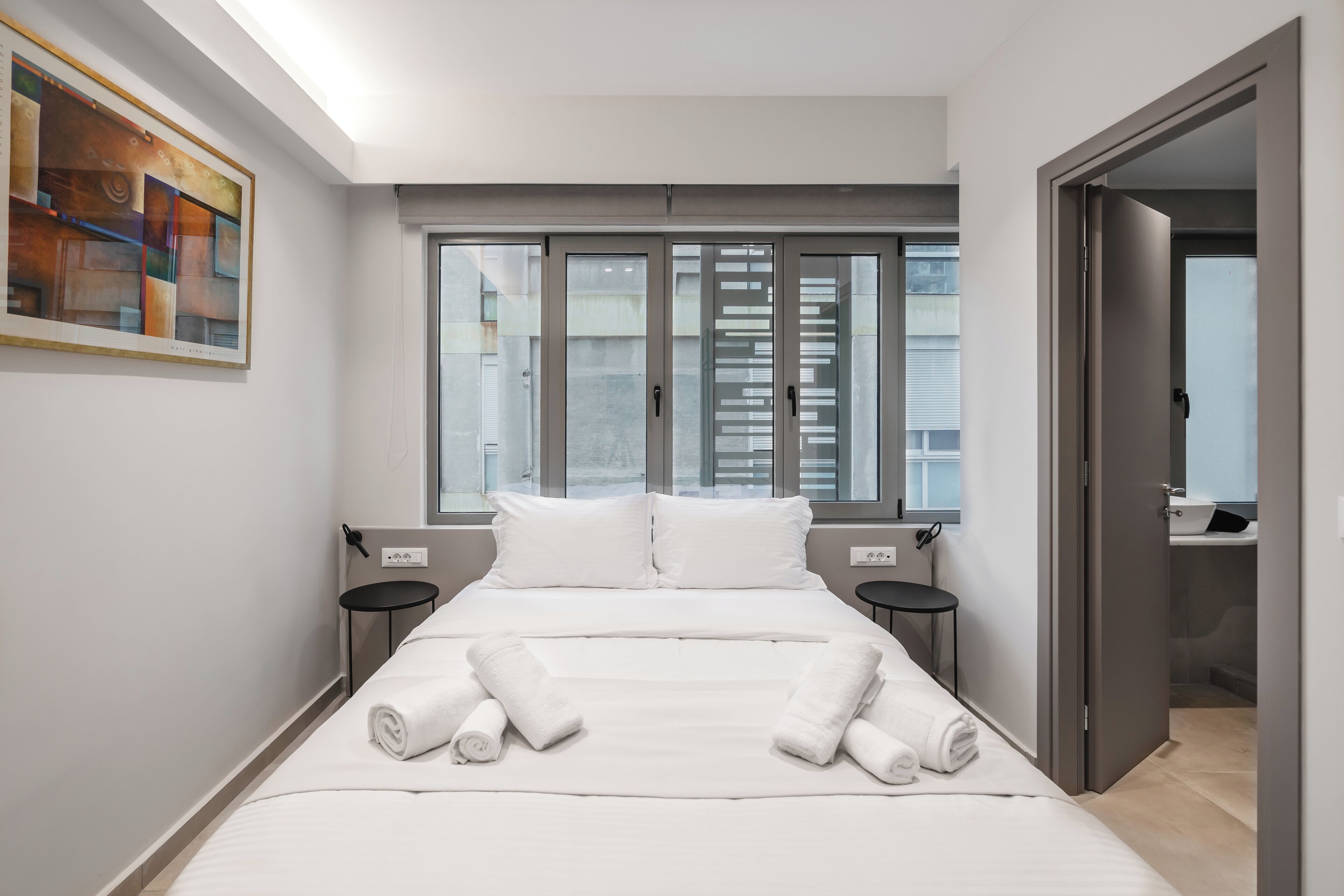 A modern hotel room with a large bed, two bedside tables, a window with blinds, and a painting on the wall.