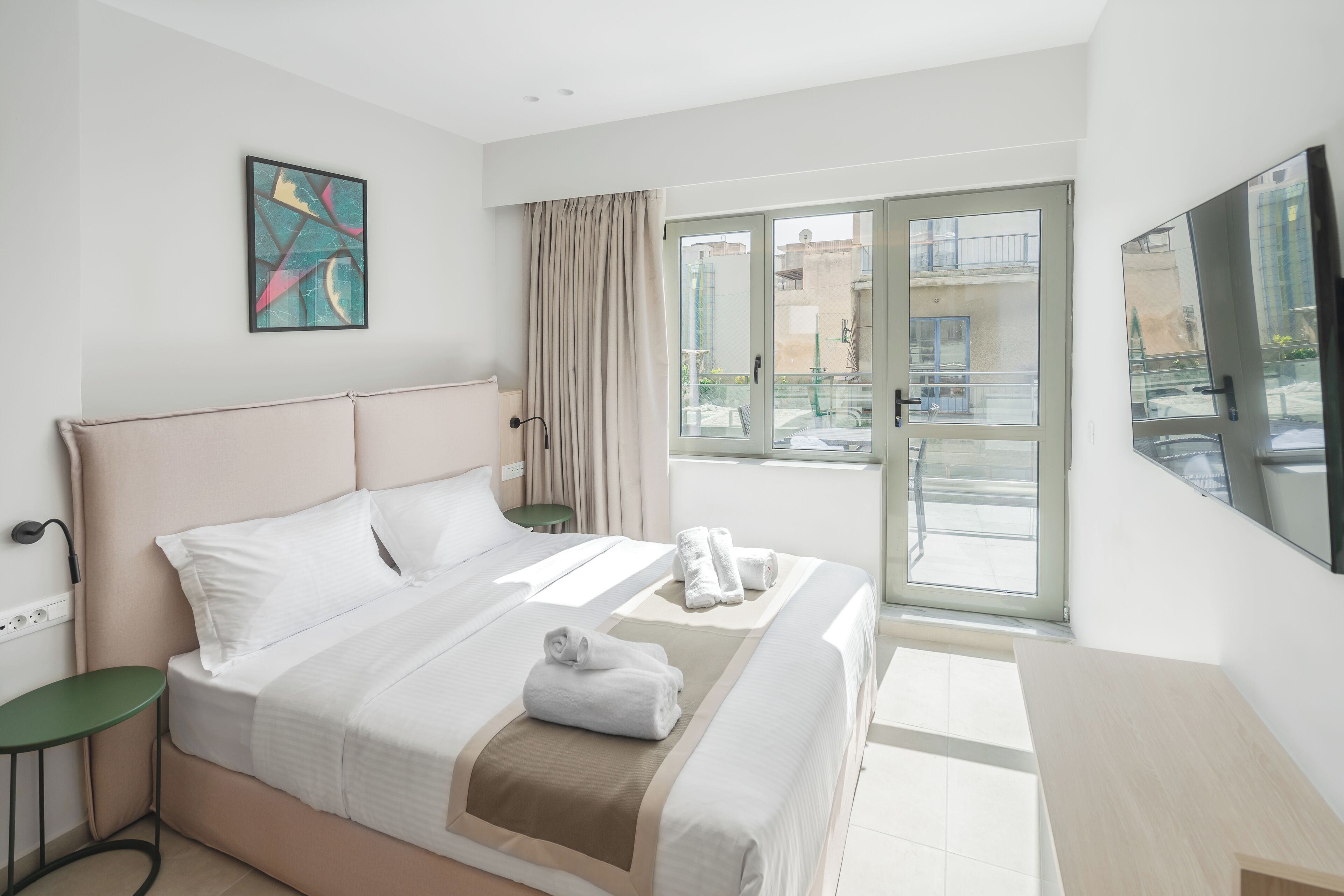 Et moderne hotelværelse med en stor seng, et lille bord, et fjernsyn og en balkon med udsigt over byen.