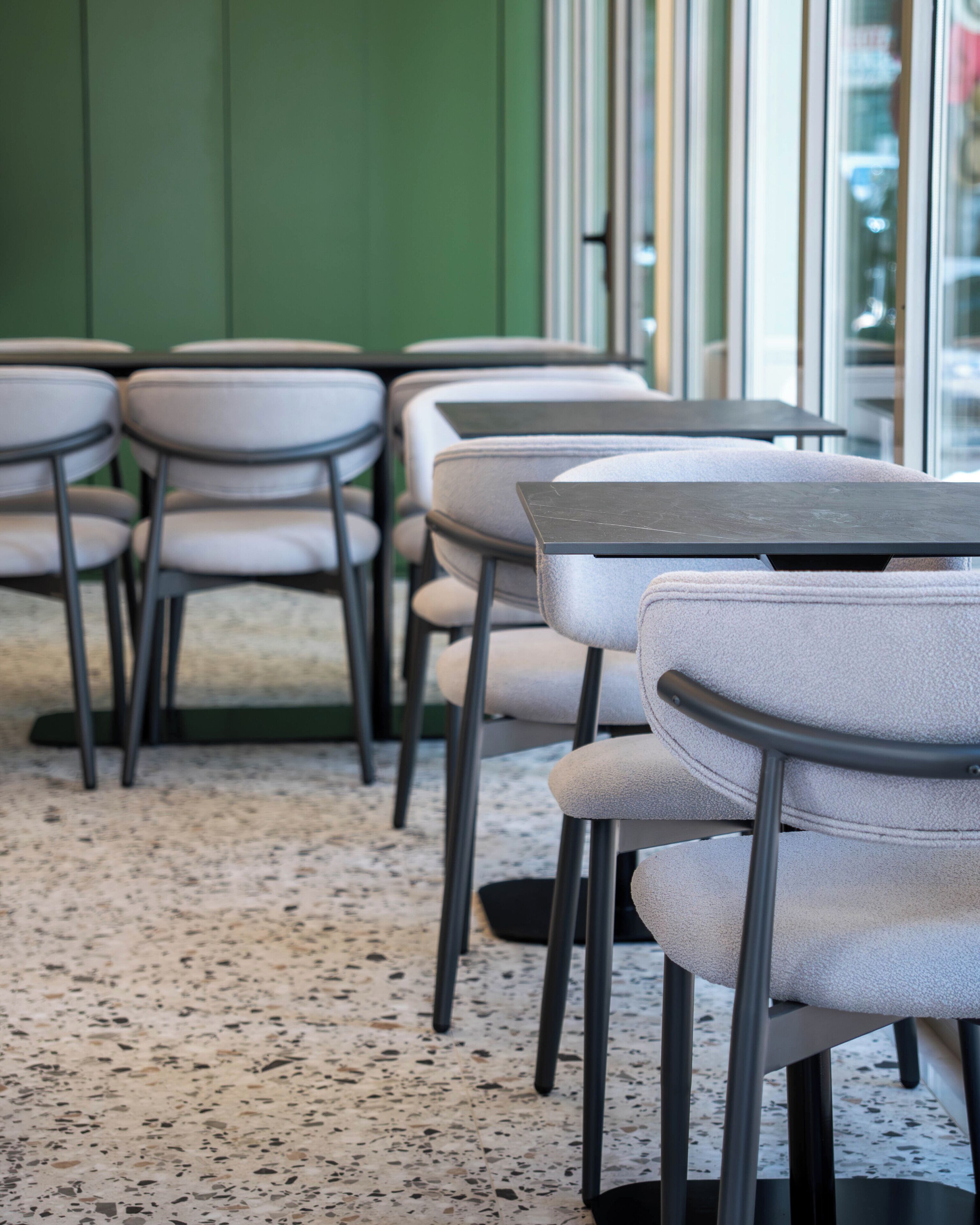 A row of tables and chairs with a terrazzo floor, green walls, and large windows.