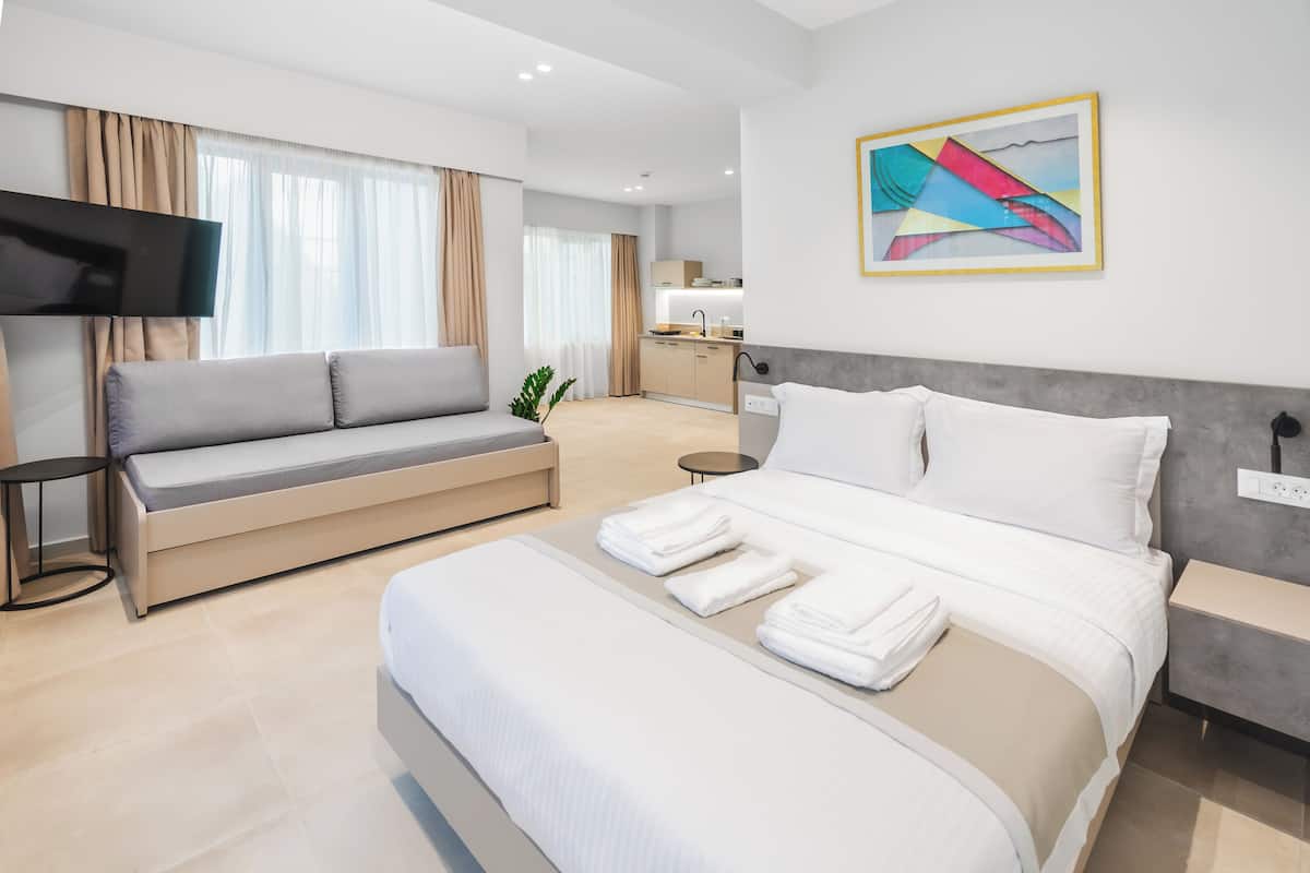 A modern hotel room with a large bed, a sofa, a flat-screen TV, and a kitchen area in the background.