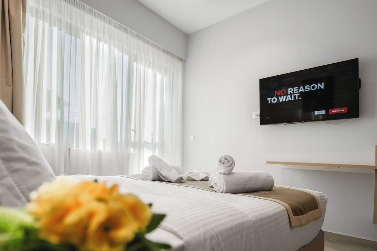 A modern hotel room with a large bed, a flat-screen TV mounted on the wall, and a window with sheer curtains.