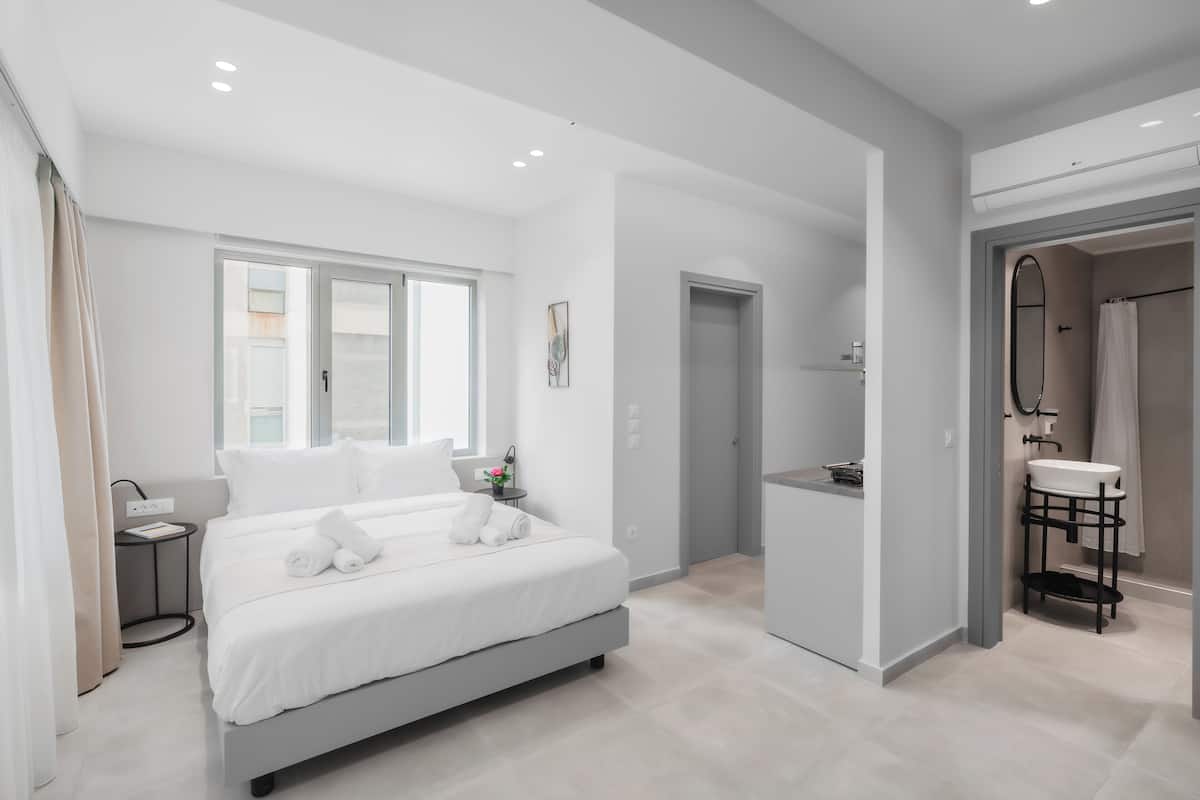 A modern hotel room with a large bed, a small bedside table, a window, and a bathroom with a sink and mirror.