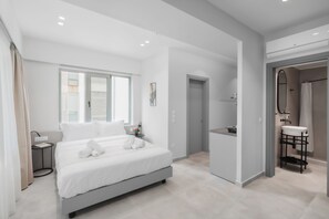 Una habitación de hotel moderna con una cama grande, una mesita de noche pequeña, una ventana y un baño con lavabo y espejo.