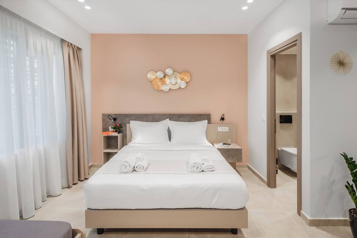 A modern hotel room with a large bed, a bedside table, a nightstand, a wall-mounted light fixture, and a bathroom visible through an open door.