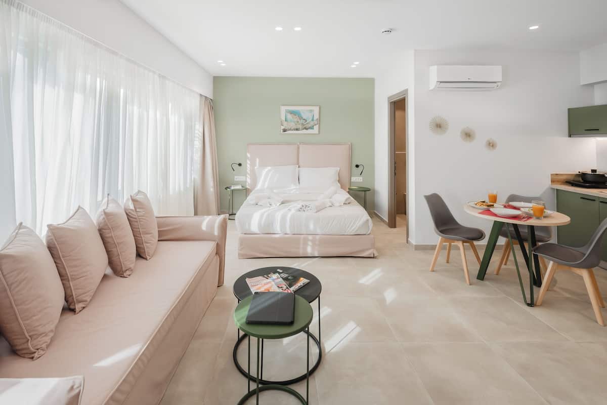 A modern hotel room with a bed, a sofa, a dining table with chairs, and a small kitchen area.