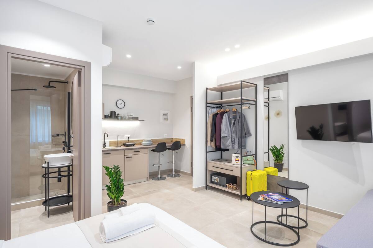 A modern hotel room with a bed, a bathroom, a kitchenette, a seating area with a TV, and a wardrobe.