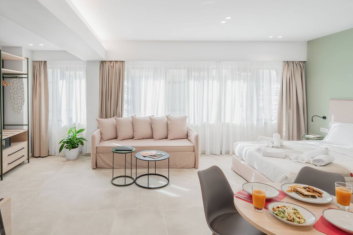 A modern hotel room with a sofa, a dining table with breakfast, a bed, and a large window with curtains.