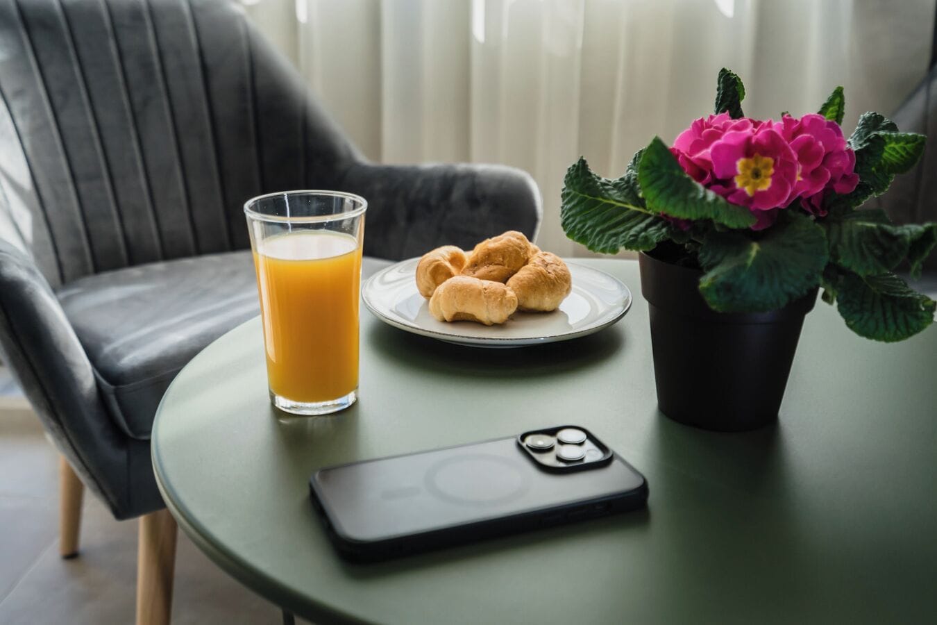 Ein Tisch mit einem Orangensaftglas, einer Croissantplatte, einem Smartphone und einem Blumentopf mit rosa Blüten.