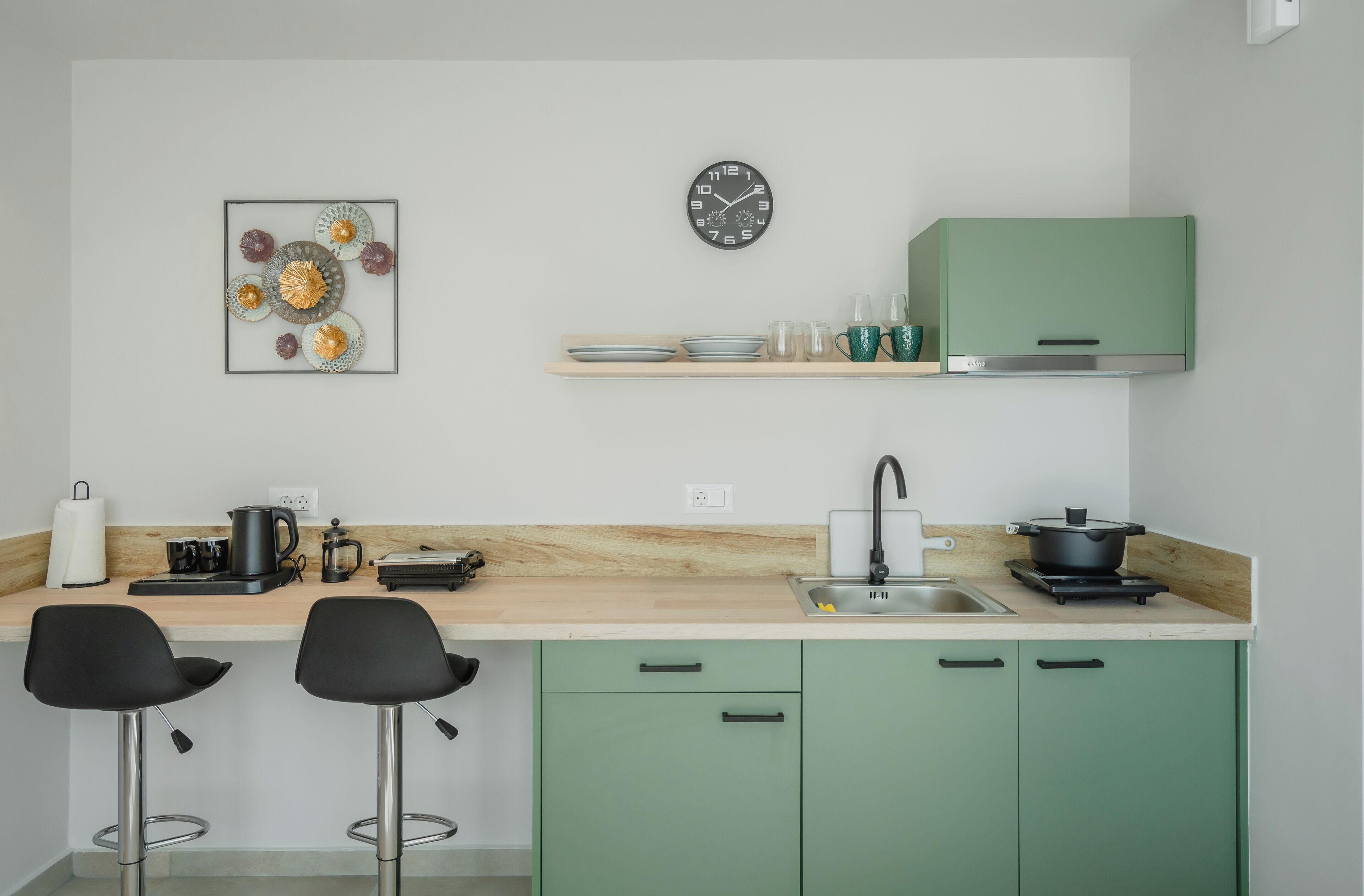 Uma cozinha moderna com armários verdes, bancada de madeira, relógio preto e um objeto decorativo na parede.