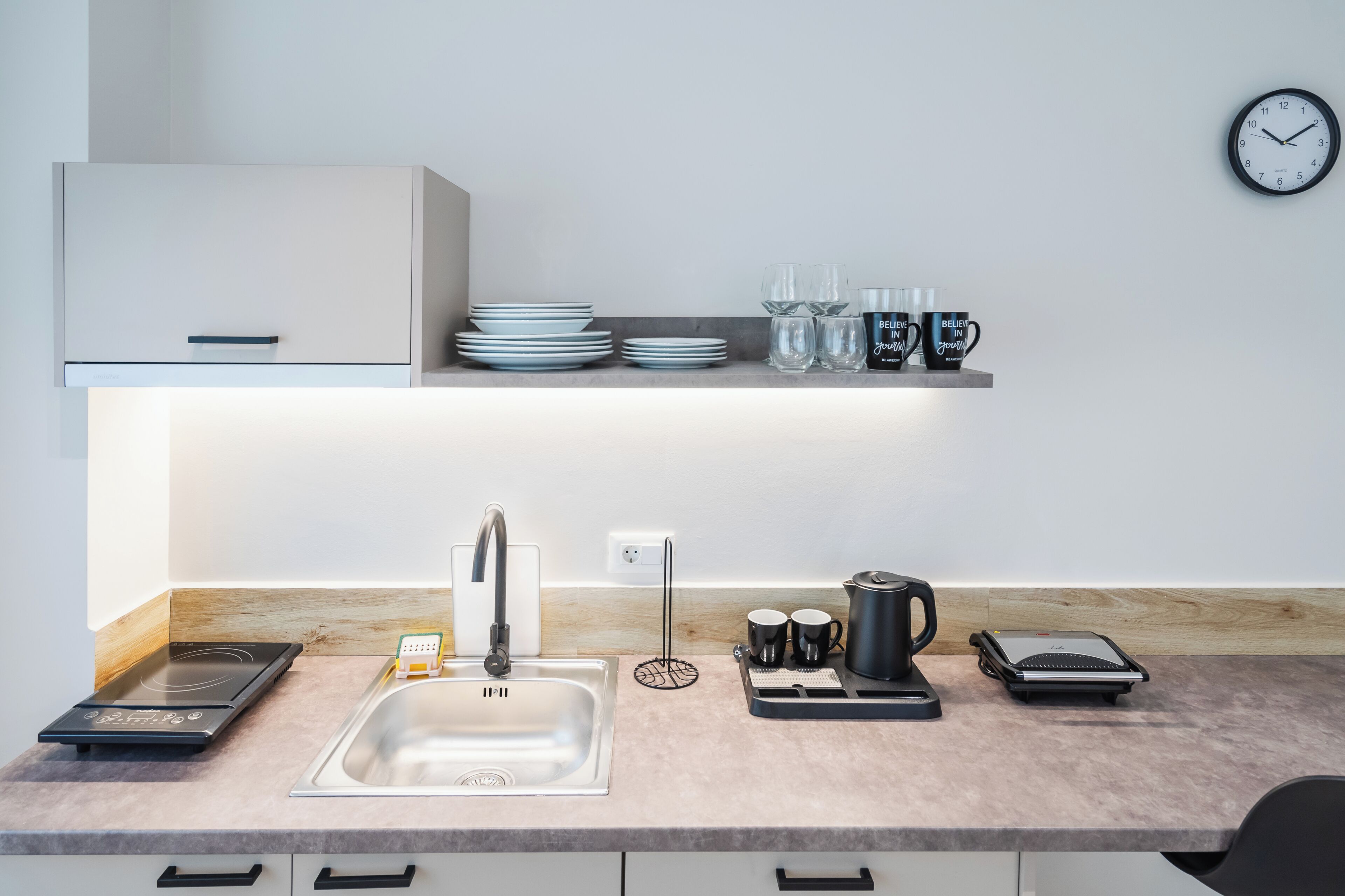 A modern kitchen with a built-in stove, sink, and countertop. There are plates, glasses, and mugs on the countertop, and a clock on the wall.