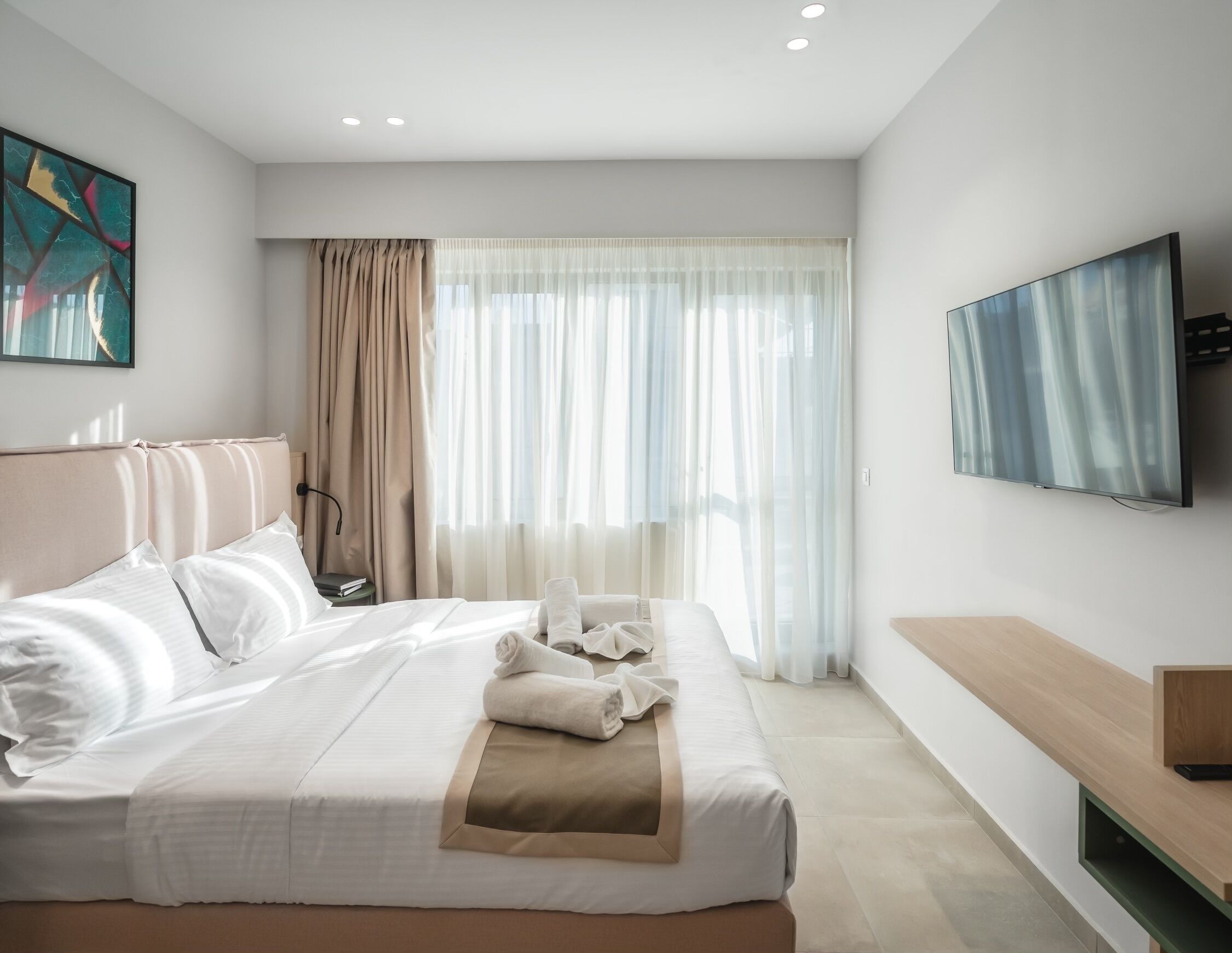 A modern hotel room with a large bed, a flat-screen TV, and a large window with sheer curtains.