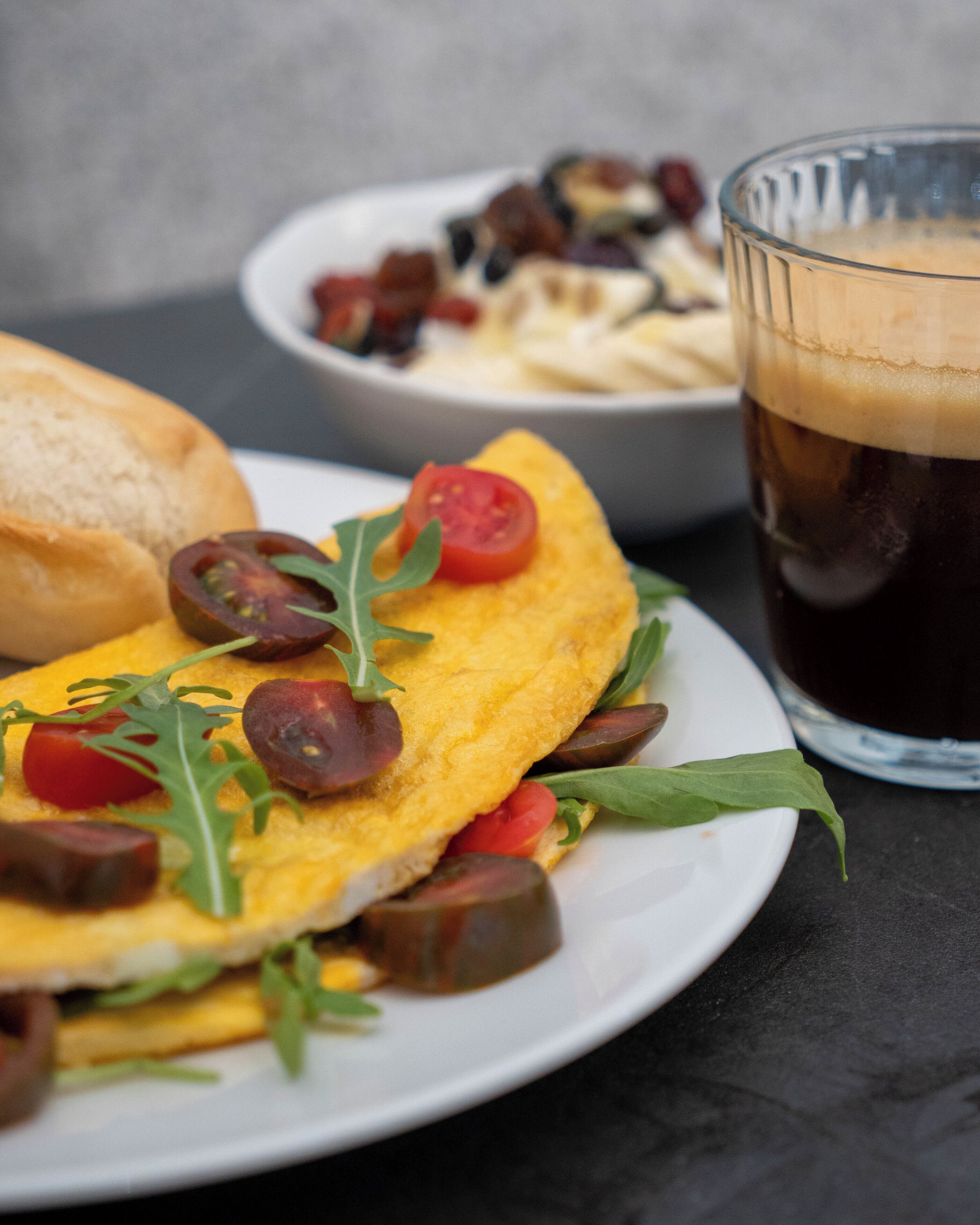 A plate with an omelette, tomatoes, and greens, a glass of dark beverage, and a bowl of fries.
