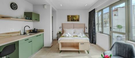 A modern hotel room with a kitchenette, a bed with a wooden headboard, a desk, and a chair.