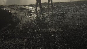 Two people standing in front of a large geyser with water spouting upwards, with the sun setting in the background.