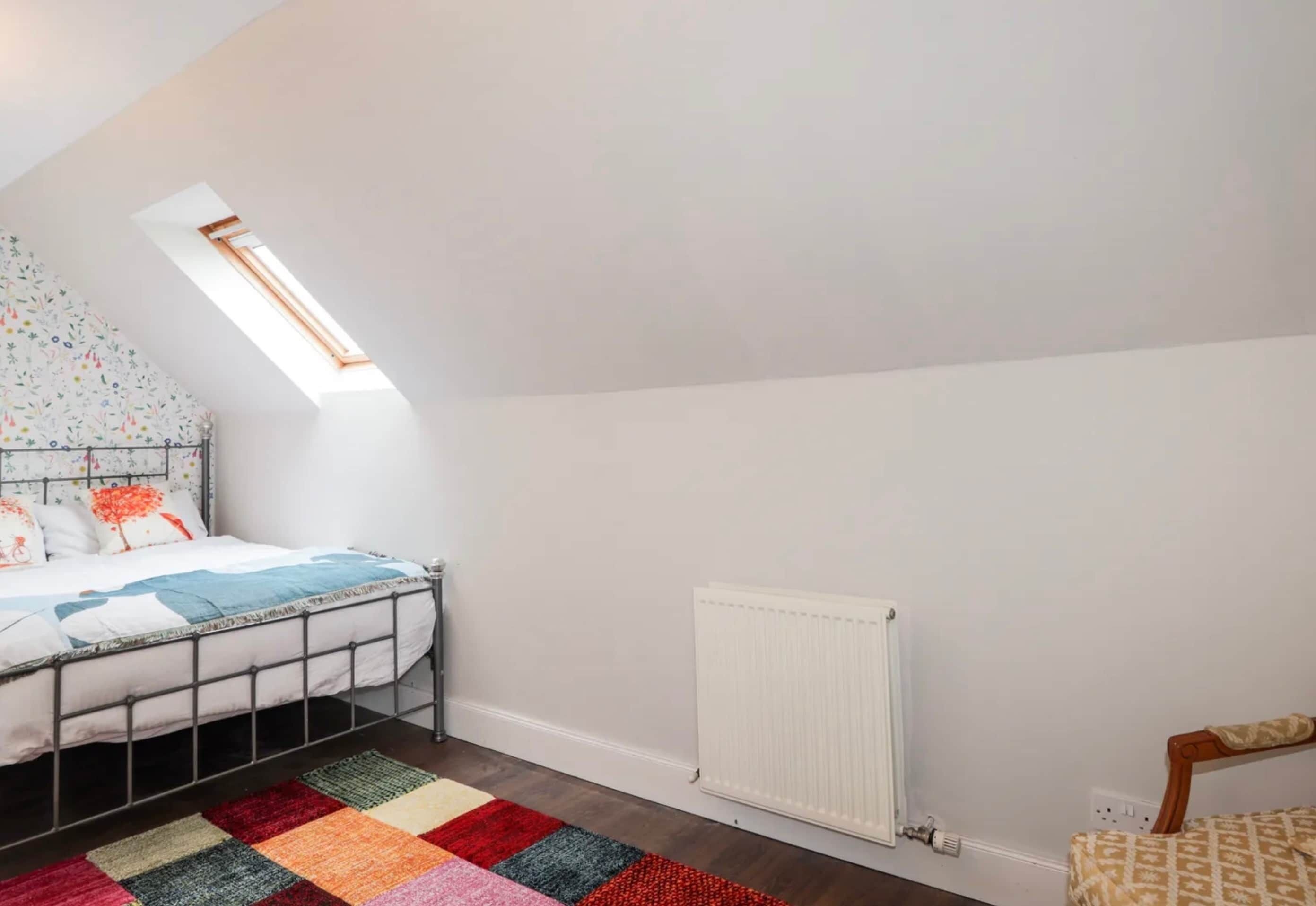 Une chambre avec un plafond en pente, un lit avec un cadre en métal, un tapis coloré, un radiateur et une petite fenêtre.