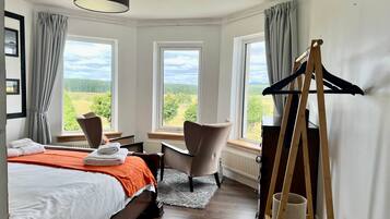 A hotel room with a bed, two chairs, a desk, a lamp, and large windows with a view of greenery.