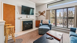 Una sala de estar con chimenea, televisor, un ventanal con vistas a árboles y un sillón azul.