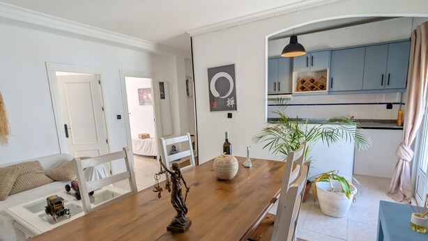 A dining area with a wooden table, chairs, and a statue centerpiece. There is a kitchen in the background with blue cabinets and a hanging light fixture. - 202 - Livingtarifa Dolce Vita (Tarifa)