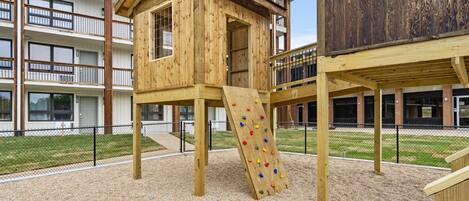 A playground with a wooden climbing structure and a slide, surrounded by a fence and a building in the background.