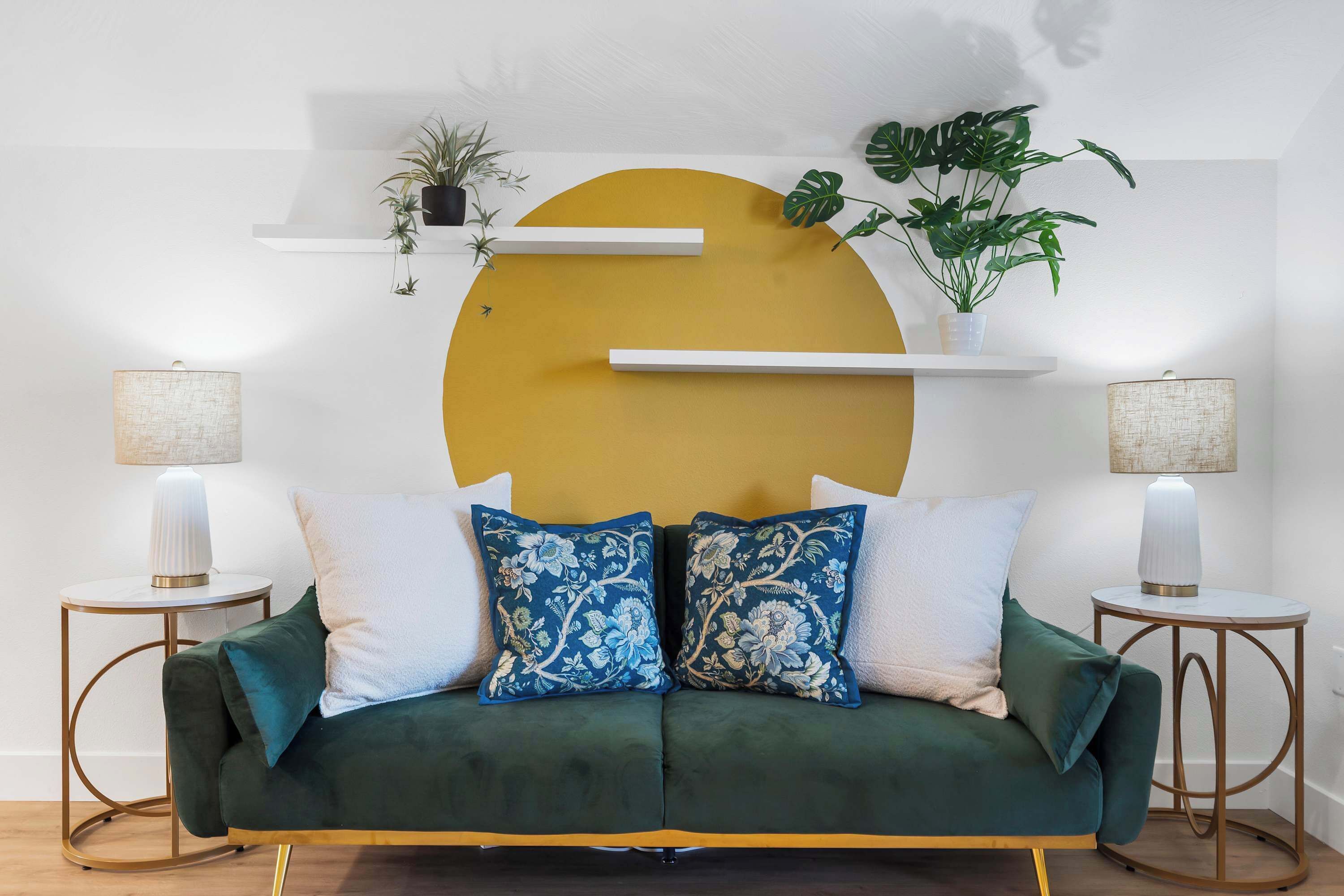 Un sofá verde con almohadones blancos y azules estampados, dos pequeñas mesitas de noche con lámparas y una decoración mural amarilla de gran tamaño.